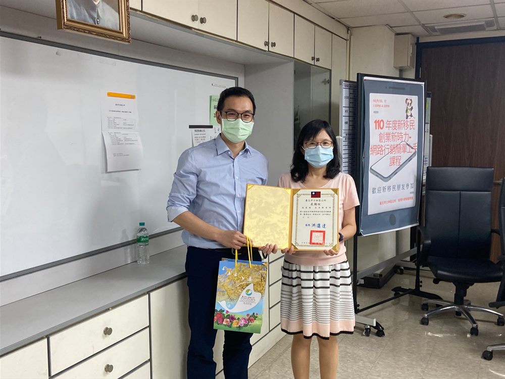 與臺北市士林區公所人員合影，手持感謝狀，旁邊是新移民網路行銷課程海報
